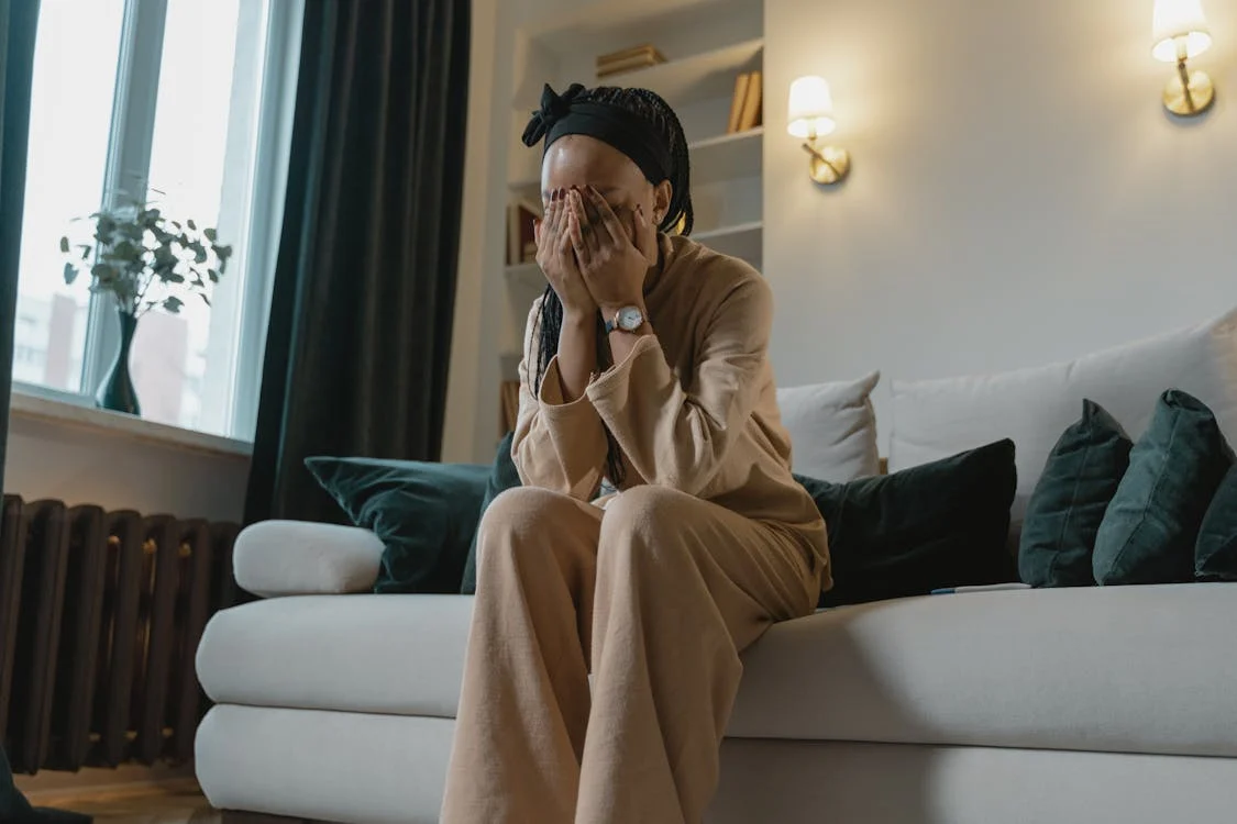 A woman sits on a couch with her head in her hands looking sad.