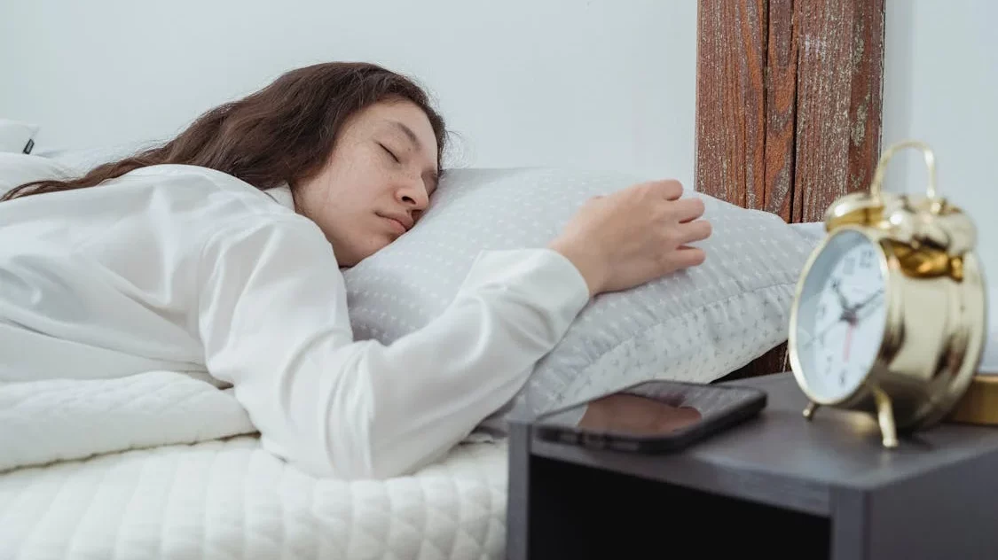 Woman laying in a bed, sleeping on her stomach while an alarm clock and cell phone sit on a bedside table next to her.