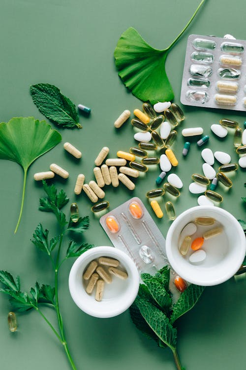 Several pills strewn about on a green surface, surrounded by various herbal leaves, plastic pill packages, and cups. The cups also have pills in them.