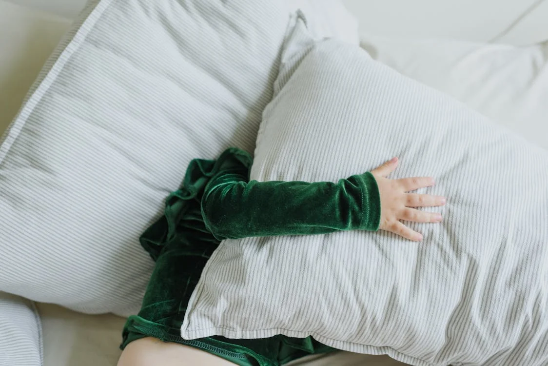 Small child laying in bed on side, holding a pillow over their head.