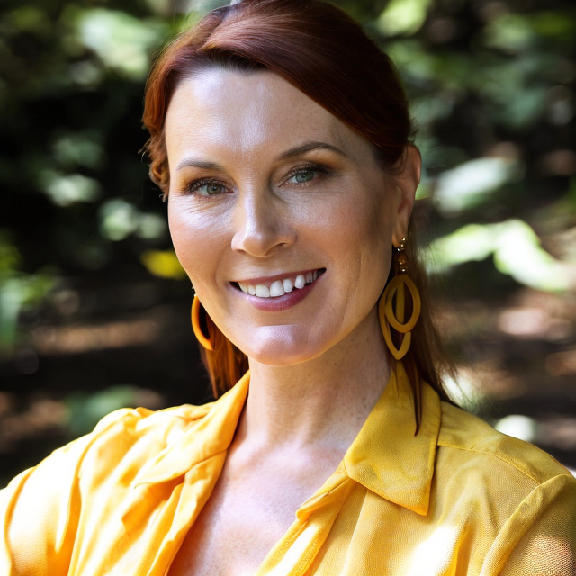 Photo of Dr. Deirdre Conroy smiling brightly in a forest, wearing brought yellow.